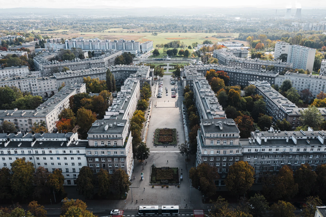 Kameralny plac otoczony symetrycznymi, wielokondygnacyjnymi kamienicami z zielenią w postaci drzew i niewielkich parkowych nasadzeń, w tle rozległy teren zielony z niską zabudową mieszkalną i lekką mgłą nad horyzontem.