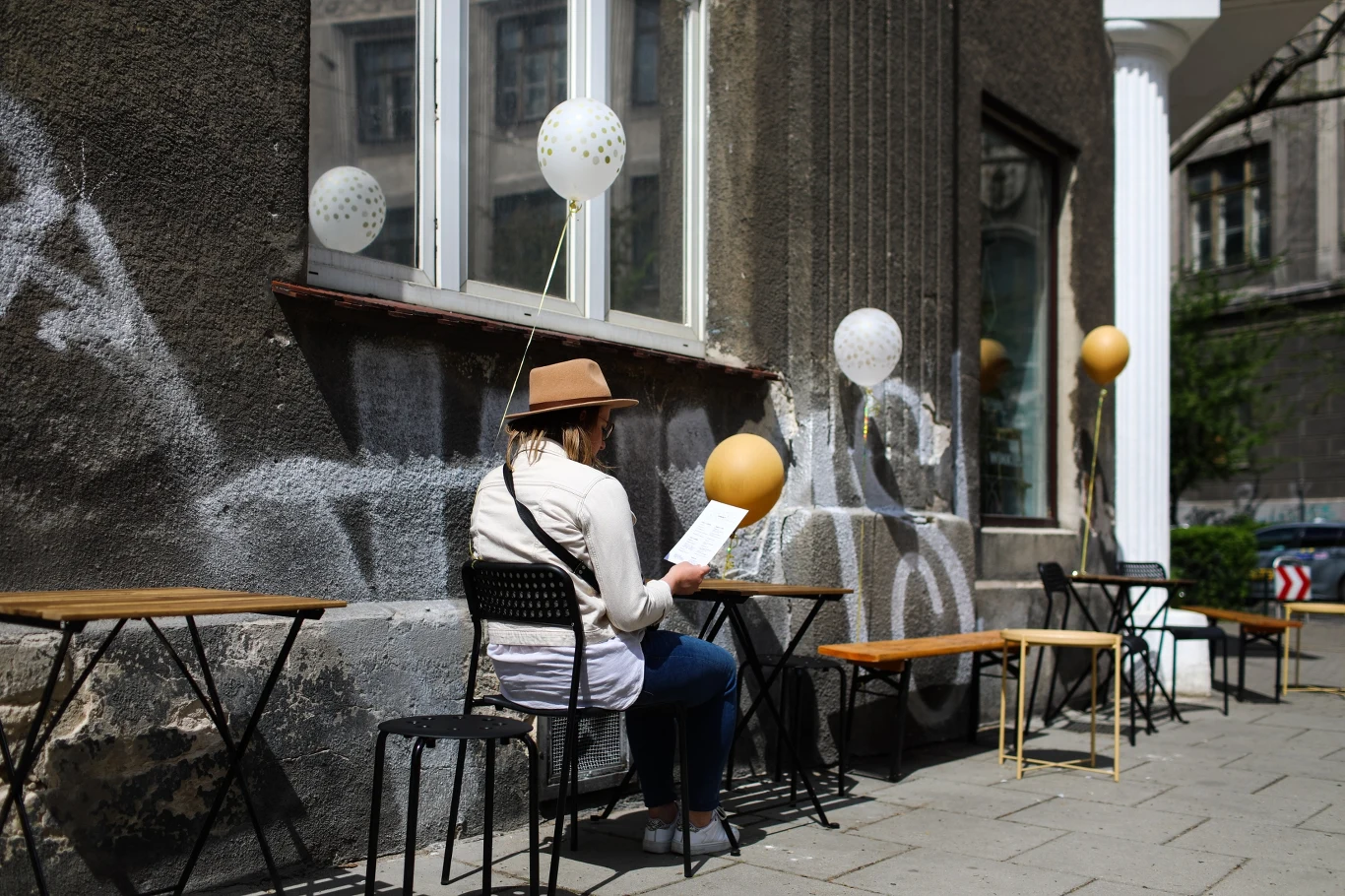 Kobieta w kapeluszu siedząca przy stoliku na zewnątrz kawiarni, trzymająca menu, otoczona balonami na tle miejskiej elewacji z graffiti.