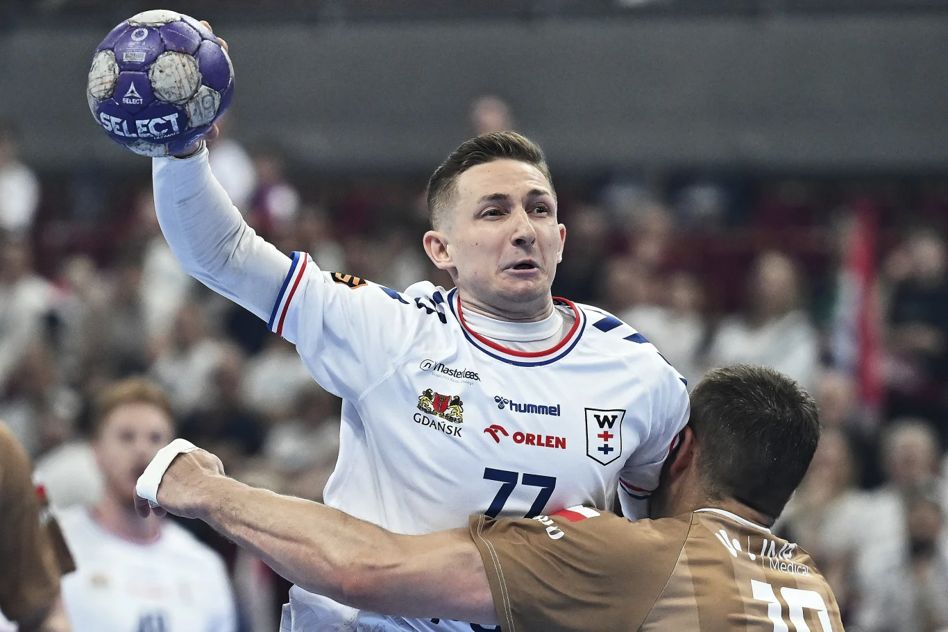 Zawodnik w białym stroju z piłką w ręce przygotowuje się do rzutu, będąc blokowanym przez przeciwnika w brązowej koszulce podczas meczu piłki ręcznej.