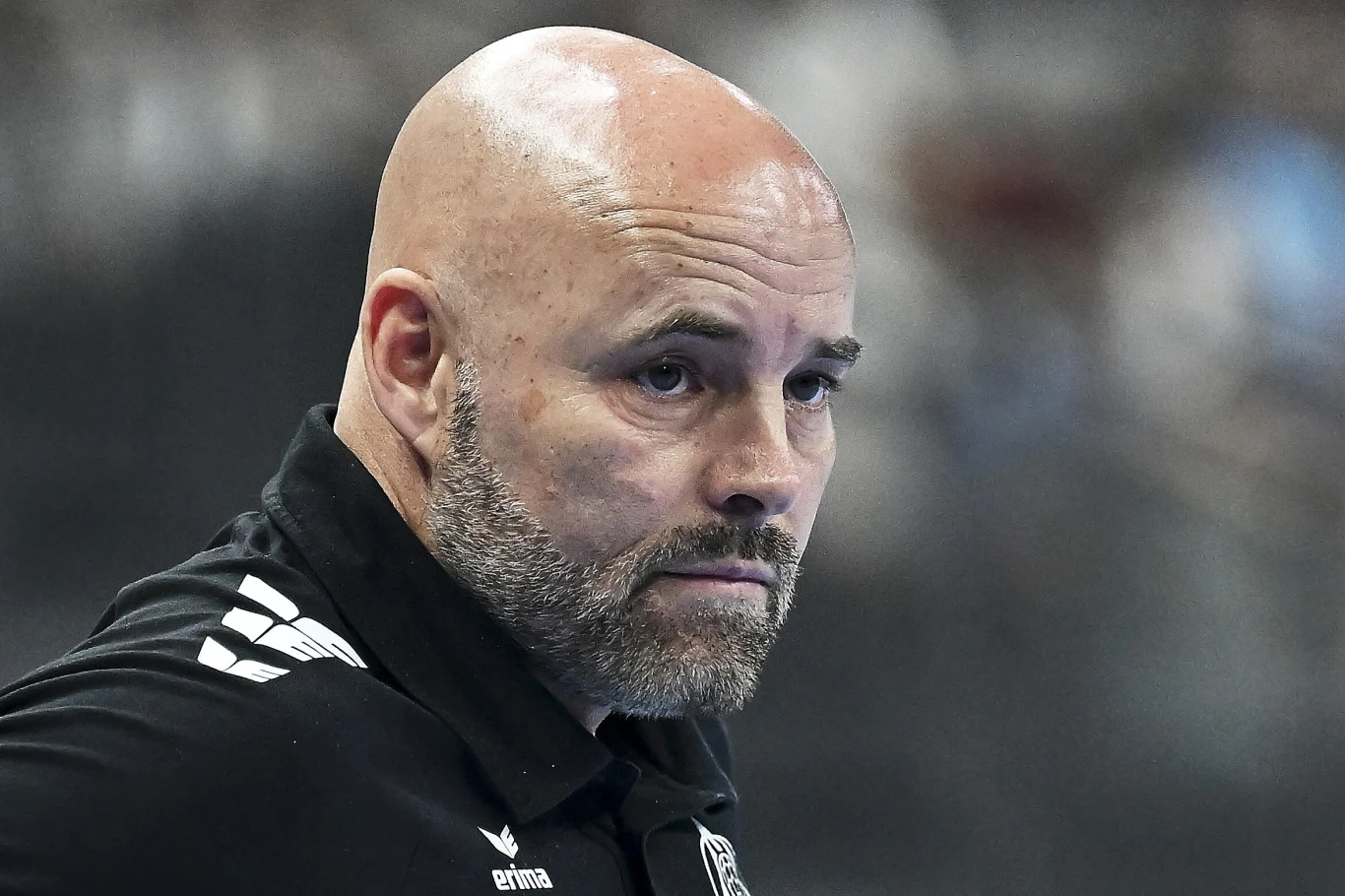 Łysy mężczyzna z krótką brodą w czarnej koszulce sportowej z widocznym logo marki Erima, skoncentrowany i patrzący w bok.