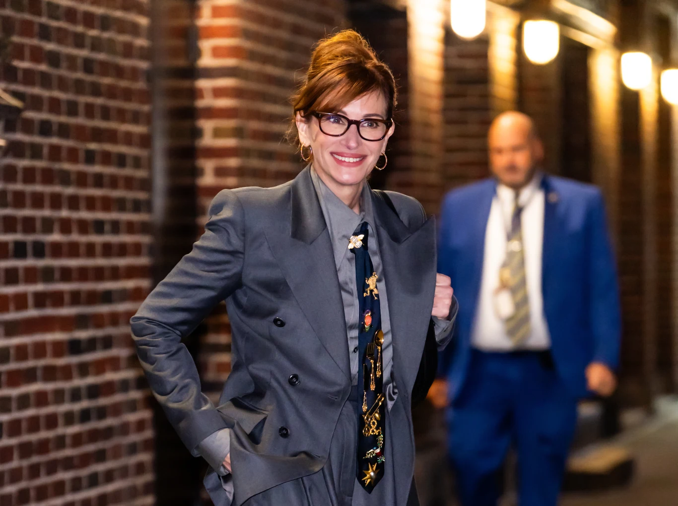 Julia Roberts zachwyca stylizacją Uśmiechnięta kobieta w szarym garniturze z wzorzystym krawatem, stojąca przed budynkiem z czerwonej cegły, w tle mężczyzna w niebieskim garniturze.