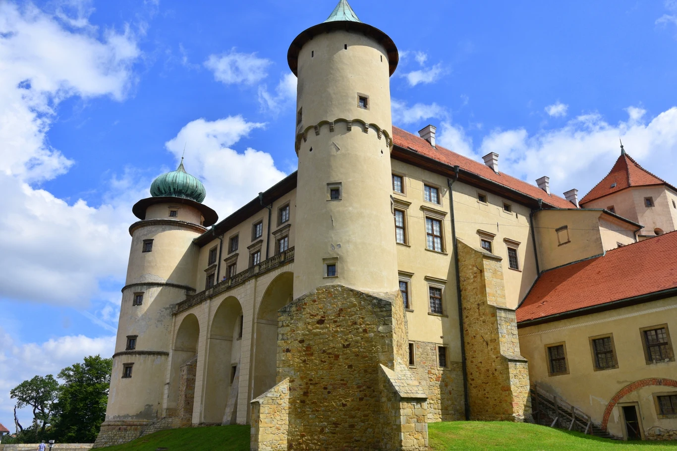 Zabytkowy zamek z okrągłymi, wysokimi wieżami i ceglanym dachem, masywne mury oraz łuki wspierające główną bryłę budynku, otoczony zielonym trawnikiem i błękitnym niebem z chmurami.