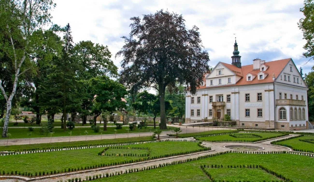 Biała rezydencja z czerwonym dachem otoczona geometrycznym ogrodem oraz licznymi drzewami w parkowej scenerii.