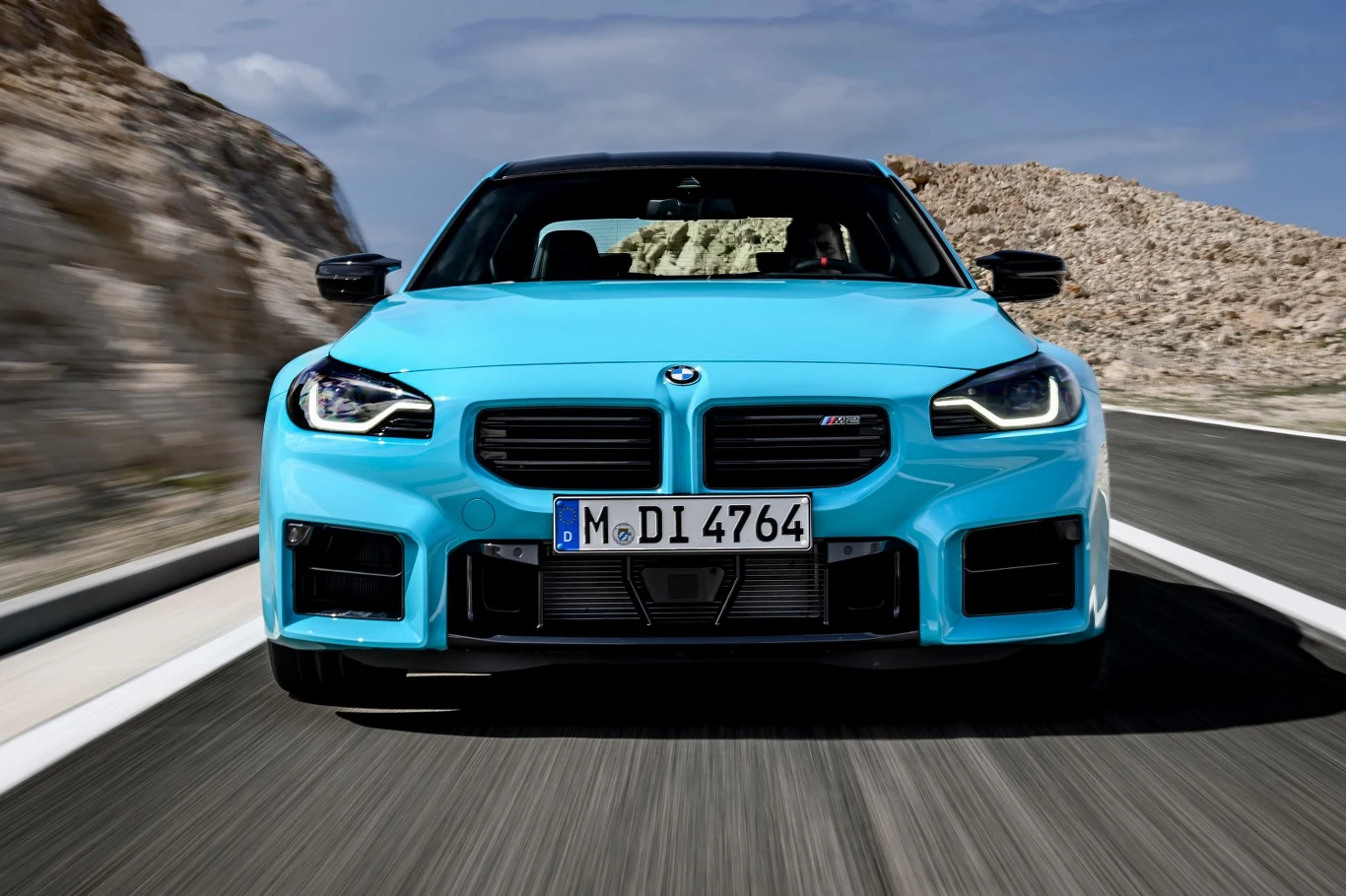 BMW M2 xDrive kosztuje w Polsce od 387 500 zł Jasnoniebieskie sportowe BMW M2 jedzie drogą pośród skalistych wzgórz, widoczne z przodu na pełnej szerokości kadru.