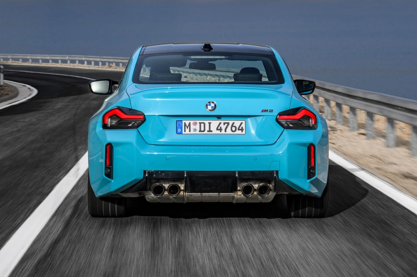 BMW M2 z napędem na 4 koła jest tańsze od M4 x Drive o 140 tys. zł Jasnoniebieskie sportowe BMW M2 z charakterystycznymi czterema końcówkami wydechu jedzie po krętej drodze, otoczonej barierkami ochronnymi.