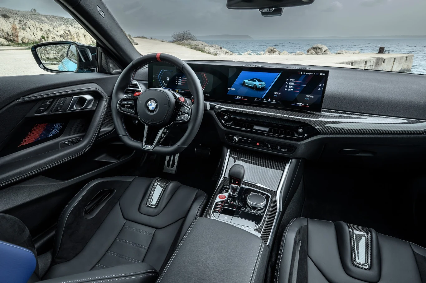 Coraz częściej słychać głosy, że BMW M2 to po prostu kompaktowa odmiana M4 Kokpit nowoczesnego samochodu BMW z cyfrowym panelem zegarów, dużym ekranem multimedialnym, skórzaną tapicerką oraz sportową kierownicą; w tle widoczna linia wybrzeża i morze.