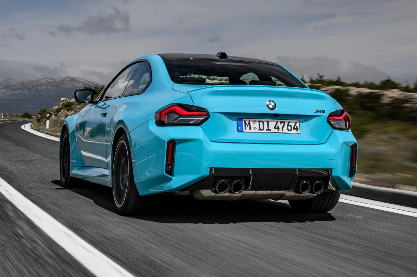 BMW M2 xDrive do 100 km/h przyspiesza w 3,7 s Jasnoniebieskie sportowe BMW M2 w dynamicznym ujęciu z tyłu, jadące po szerokiej, asfaltowej drodze z wyraźnymi białymi liniami, otoczone górskim krajobrazem.