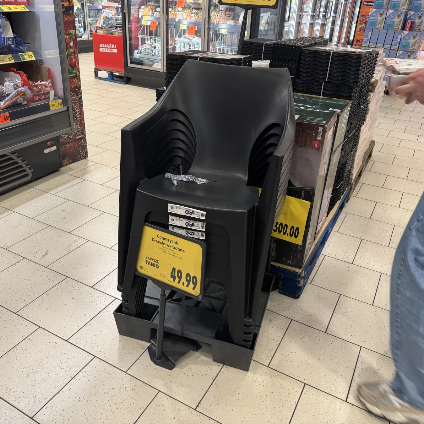 Czarny plastikowy fotel ogrodowy ustawiony na stojaku w sklepie, z wyeksponowaną dużą żółtą ceną 49,99 i kilkoma zawieszonymi etykietami na siedzisku.