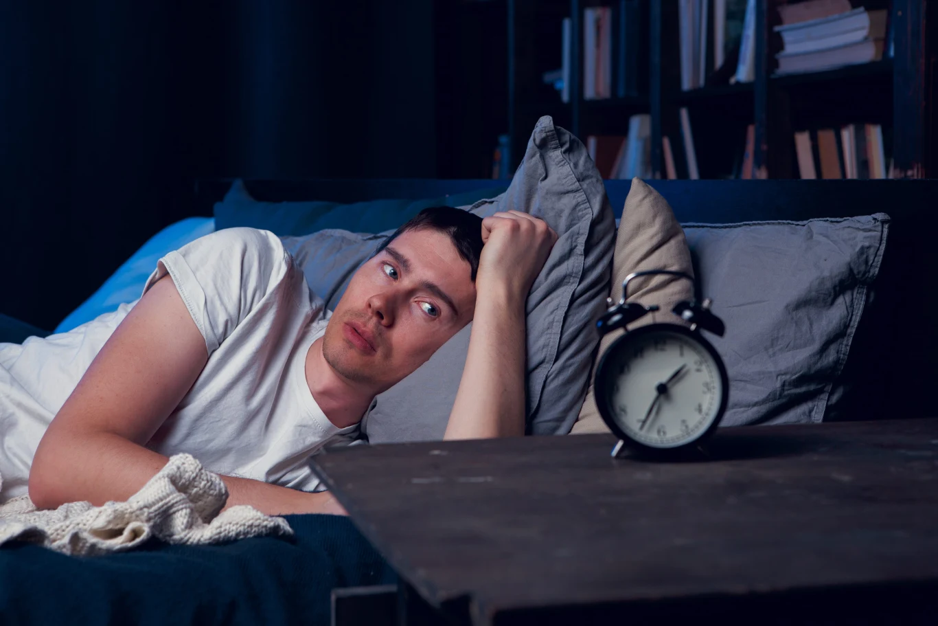 U sów naturalna senność pojawia się dopiero między godziną 2:00 a 6:00 rano. Mężczyzna leżący na łóżku z głową opartą na ręce, patrzący z niepokojem w stronę budzika ustawionego na stoliku nocnym.