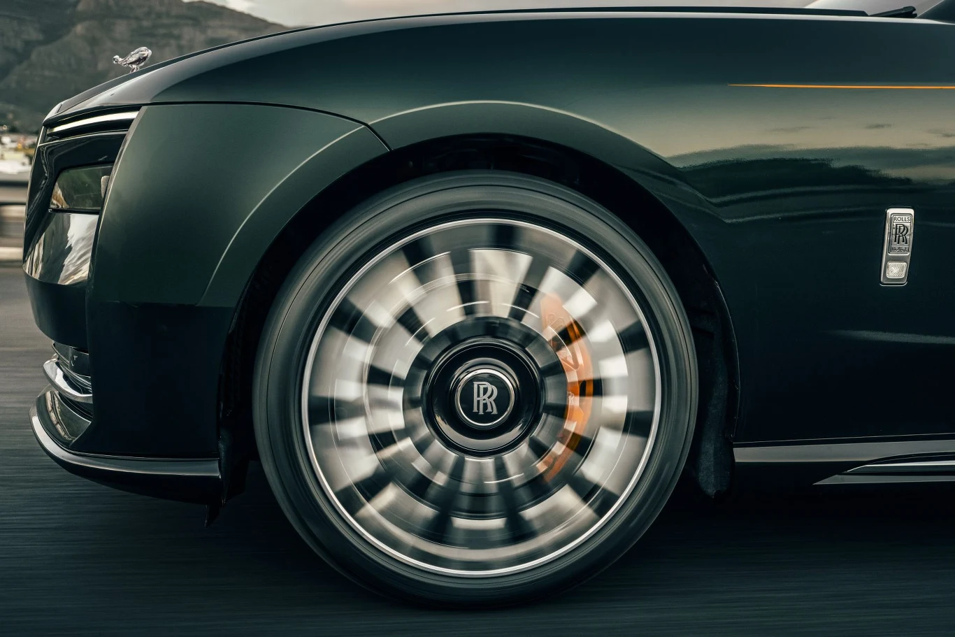 Zbliżenie na obracające się koło luksusowego samochodu marki Rolls-Royce jadącego po drodze, z wyraźnie widocznym emblematem producenta na środku felgi.