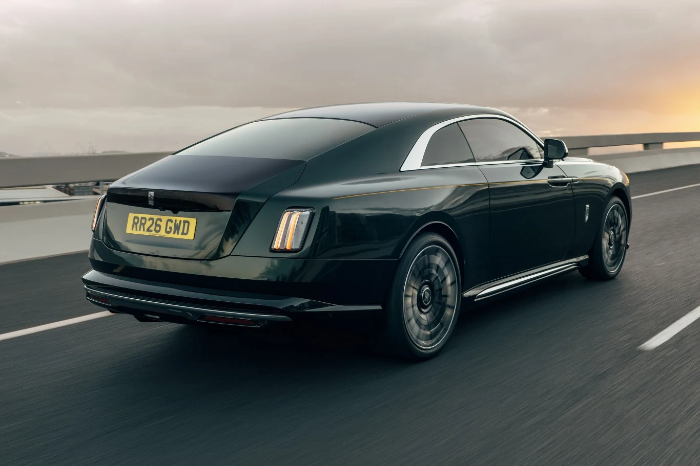 Czarny luksusowy samochód coupe marki Rolls-Royce jedzie po pustej, asfaltowej drodze przy zachmurzonym niebie, ujęcie z tyłu w dynamicznym ruchu.