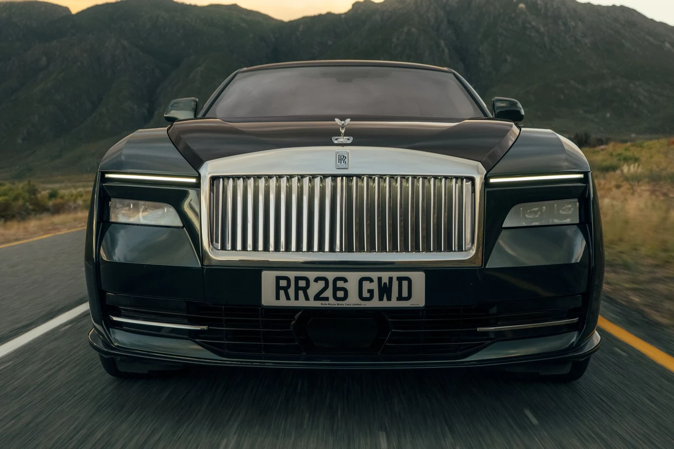 Luksusowy czarny Rolls-Royce z imponującą chromowaną atrapą chłodnicy i charakterystycznym emblematem Spirit of Ecstasy jadący drogą przy górzystym krajobrazie.