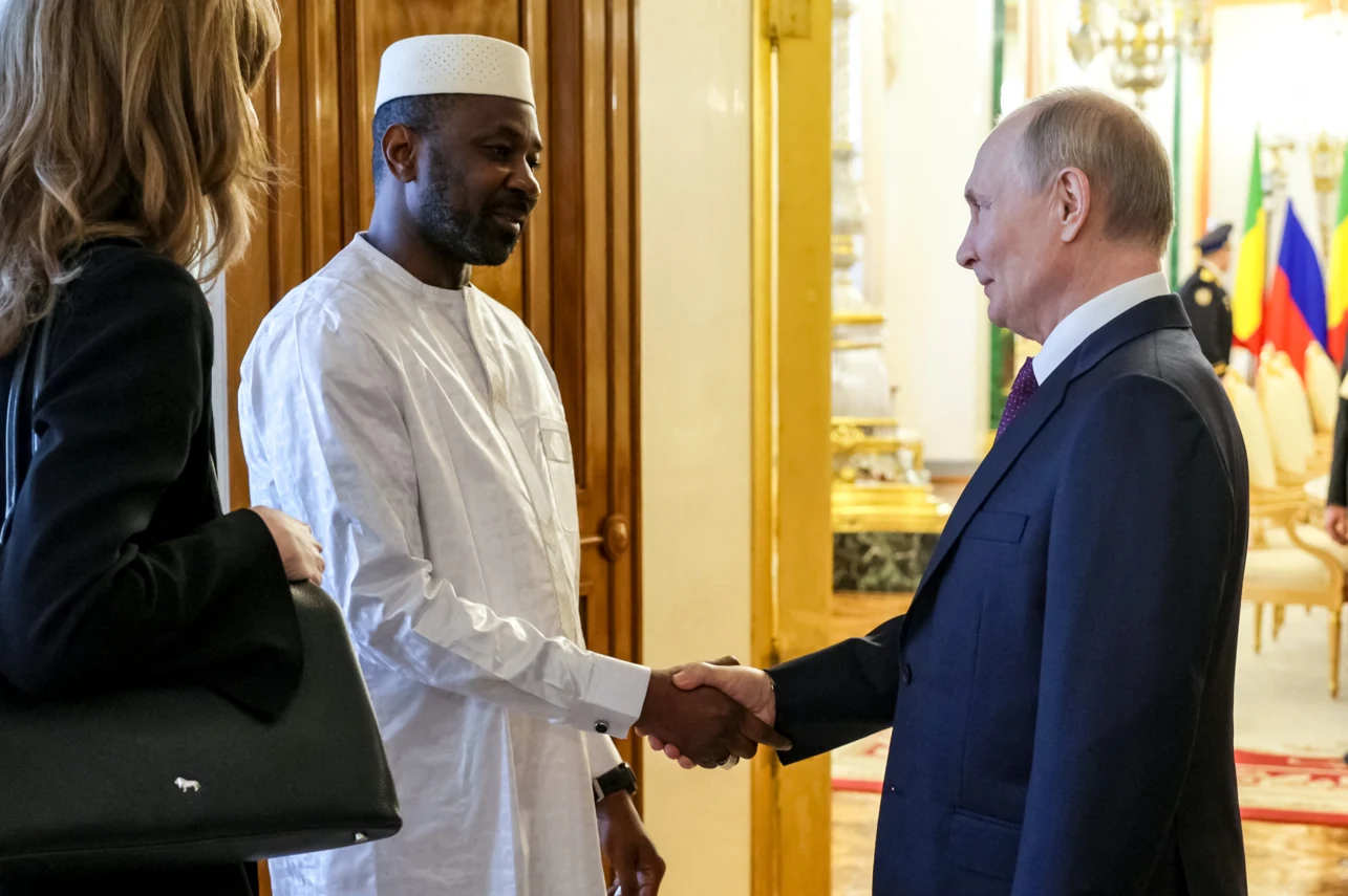 Dwóch mężczyzn wymienia uścisk dłoni w eleganckim wnętrzu, jeden z nich ubrany w tradycyjny biały strój i czapkę, drugi w formalny garnitur; w tle widoczne flagi państwowe.