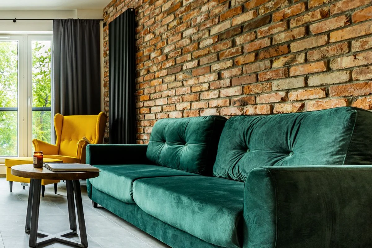 https://www.e-ceglanafantazja.pl/ Zielona, welurowa sofa ustawiona na tle ceglanej ściany, obok żółty fotel przy dużych oknach z zasłonami, pośrodku niski stolik z kawą.