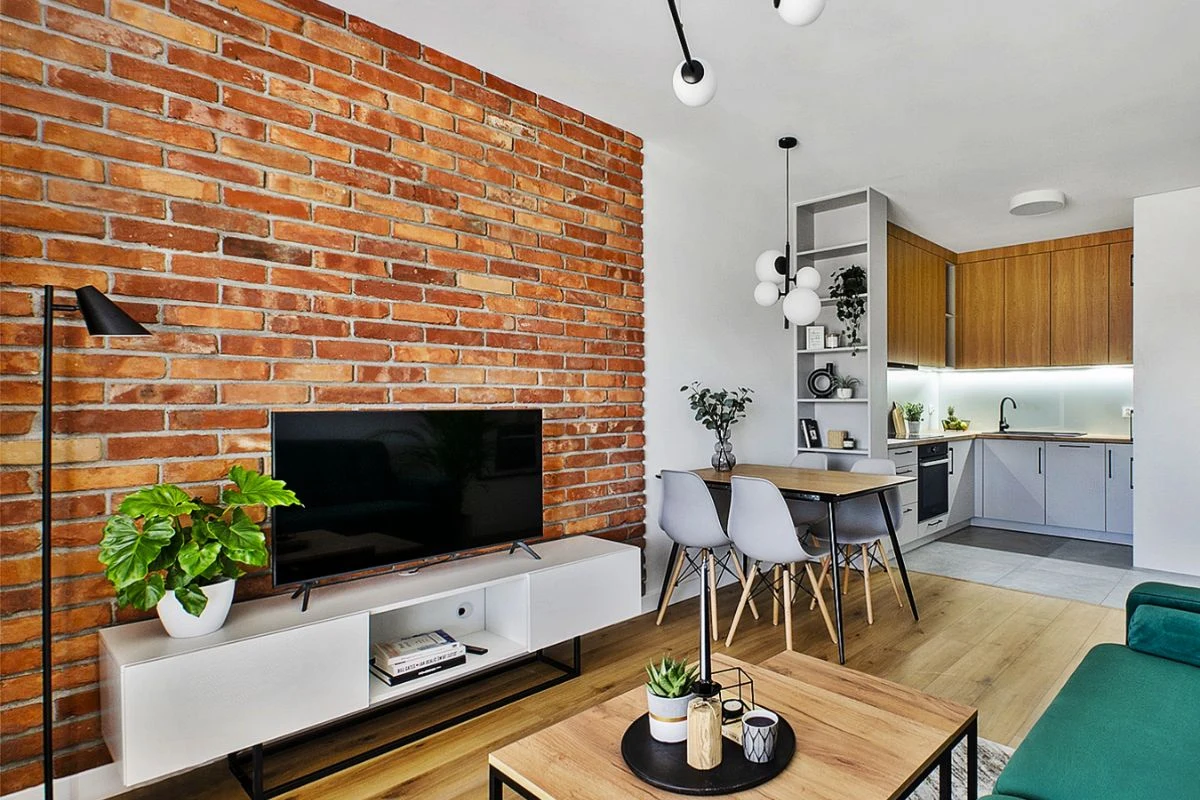 https://www.e-ceglanafantazja.pl/ Salon z ceglaną ścianą, nowoczesnym telewizorem na białej szafce, zieloną kanapą, stolikiem z ozdobami oraz aneksem kuchennym w drewnie i szarościach, obok jadalni z czterema krzesłami.