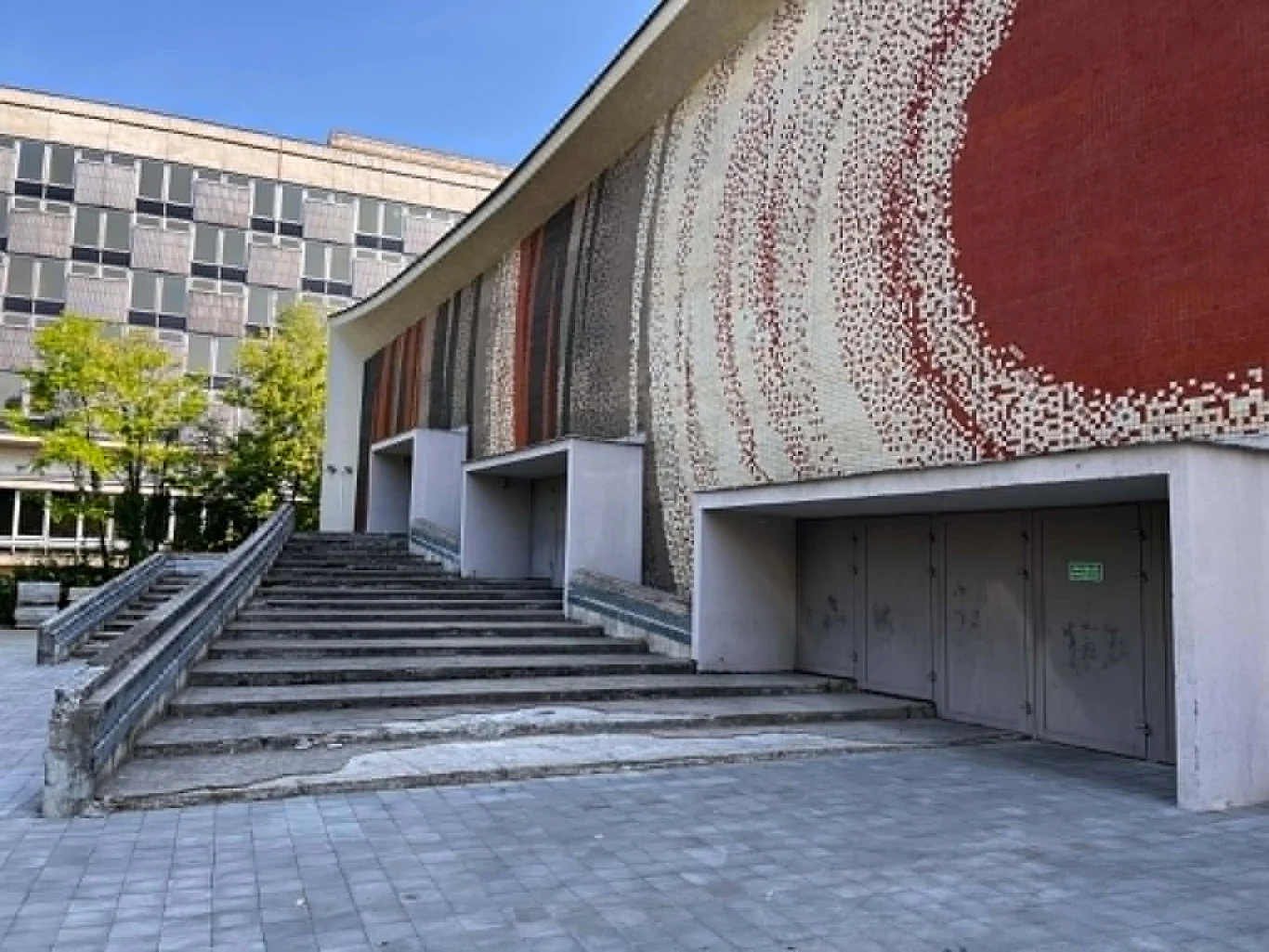 Szerokie schody prowadzące do nowoczesnego budynku z charakterystyczną, mozaikową fasadą w odcieniach czerwieni, beżu i brązu, ozdobioną geometrycznymi wzorami.