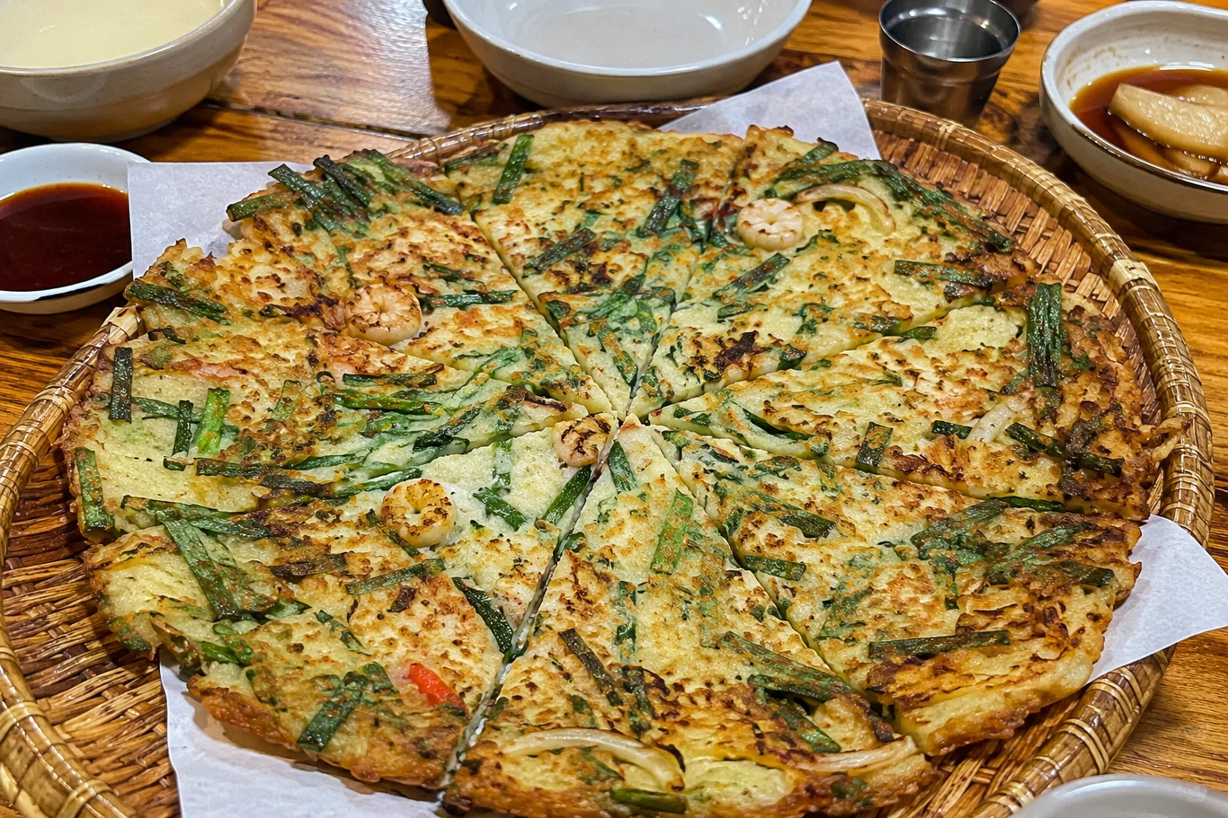 Pajeon można podawać z różnymi dodatkami Duży, pokrojony w trójkątne kawałki placek z widocznymi kawałkami owoców morza, zieloną cebulką i warzywami, podany na okrągłym koszu wyłożonym papierem, z dodatkiem miseczek z sosami i dodatkami wokół.