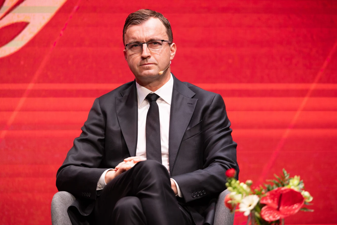 Mężczyzna w garniturze i okularach siedzący na krześle na tle czerwonej ściany, obok dekoracyjna kompozycja kwiatowa.