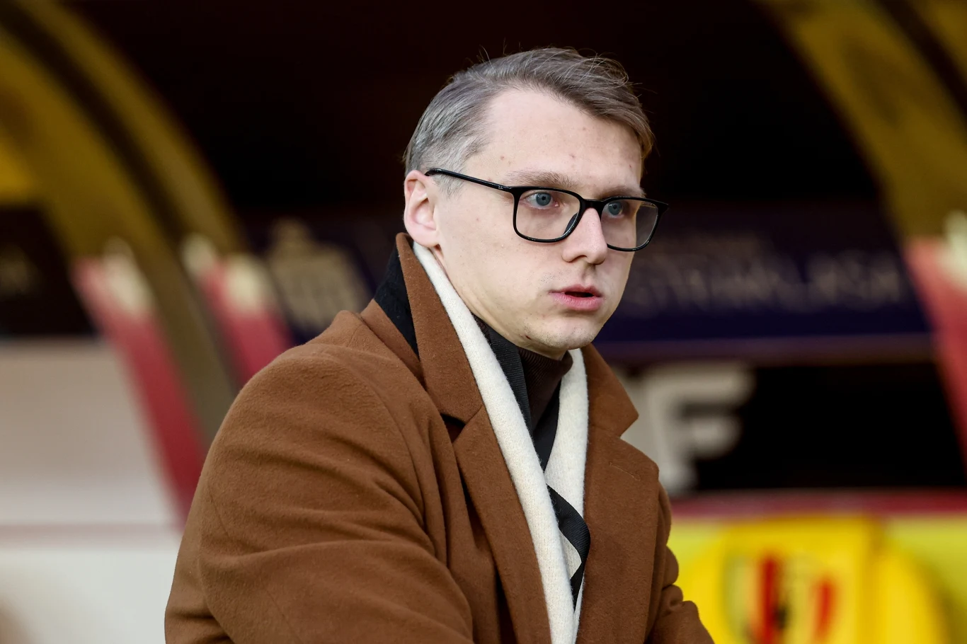 Mężczyzna w okularach i brązowym płaszczu z kremowym szalem, siedzący w miejscu przypominającym stadion lub trybuny.