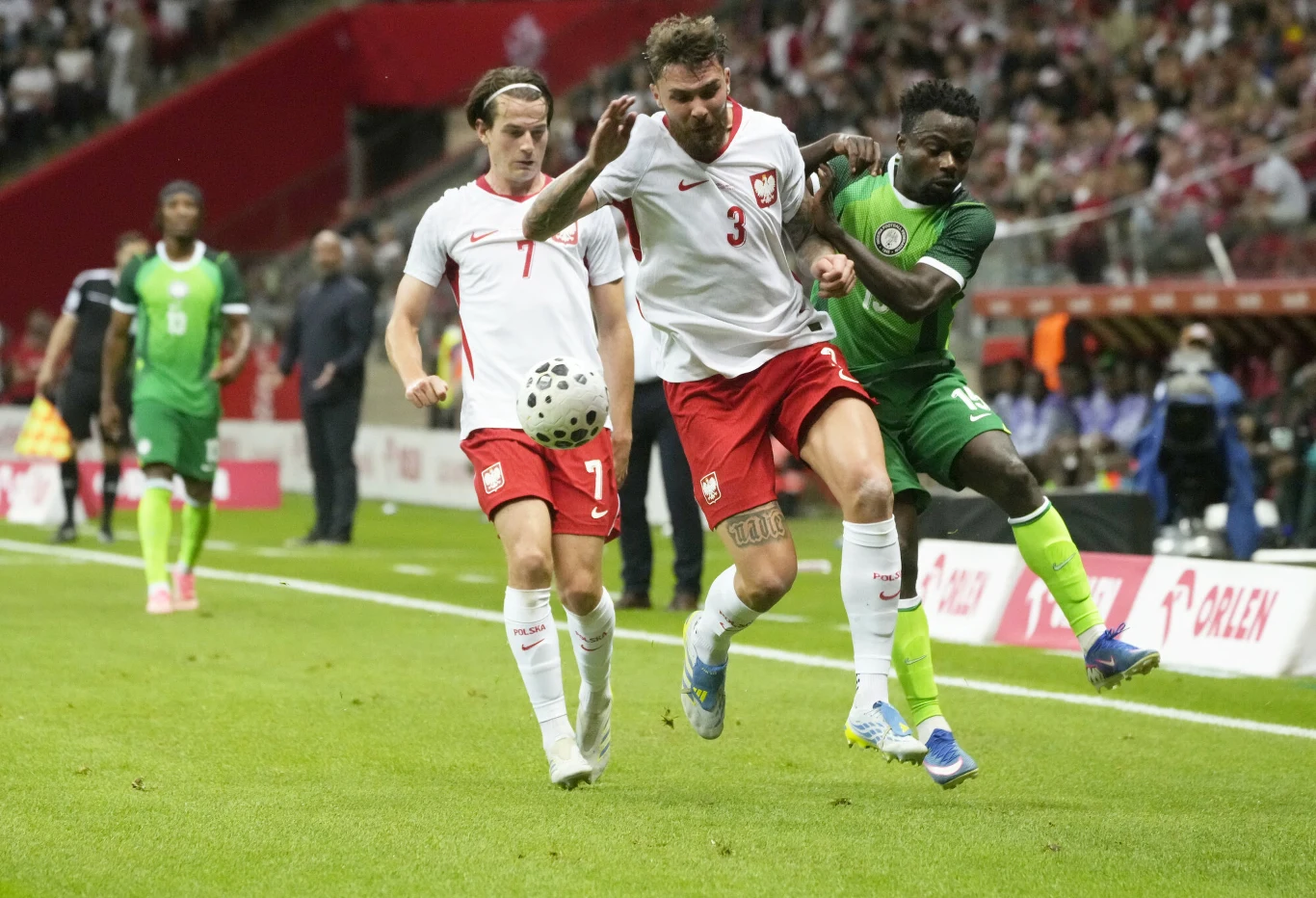 Polski piłkarz walczy o piłkę z zawodnikiem w zielonym stroju podczas meczu na stadionie pełnym kibiców, a obok nich biegnie kolejny piłkarz w białej koszulce.