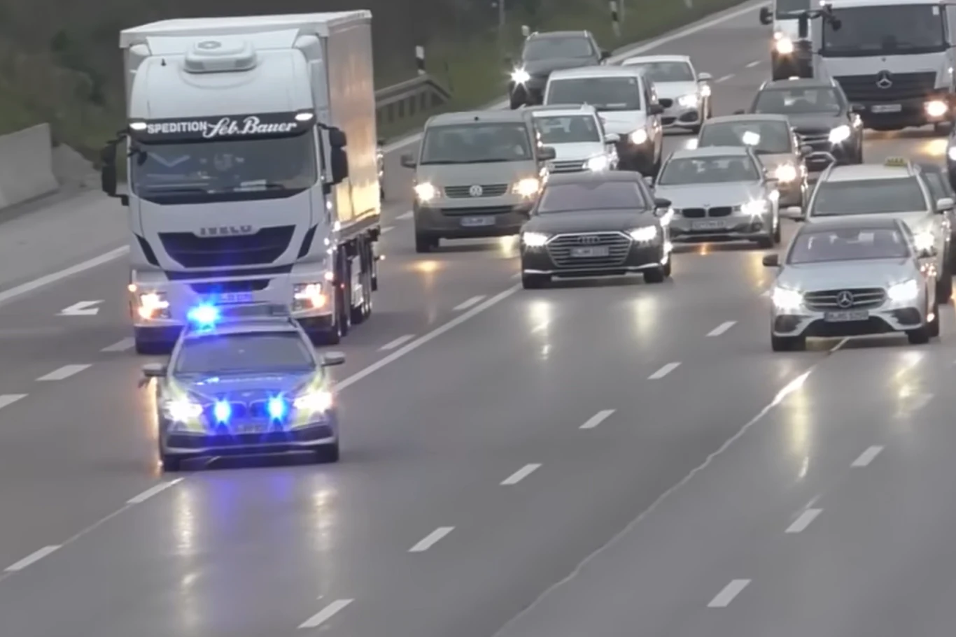Jadać zygzakiem policjanci wyhamowują ruch. / YouTube - EnfoyFireFighting - Inernational Emergency Response Videos Radiowóz policyjny z włączonymi sygnałami świetlnymi jedzie lewym pasem autostrady, prowadząc kolumnę samochodów oraz ciężarówkę w intensywnym ruchu ulicznym.