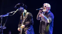 Dwóch artystów na scenie podczas koncertu – pierwszy z nich nosi maskę i kostium z czarnymi piórami, gra na gitarze elektrycznej, drugi śpiewa do mikrofonu, ubrany w czarną koszulkę i okulary przeciwsłoneczne o jasnych oprawkach.