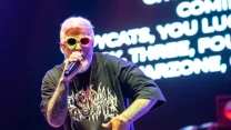 Brodaty artysta w kolorowych okularach i czarnej koszulce z nadrukiem rapuje do mikrofonu na scenie, w tle widoczny fragment bębnów i podświetlony ekran z tekstem.