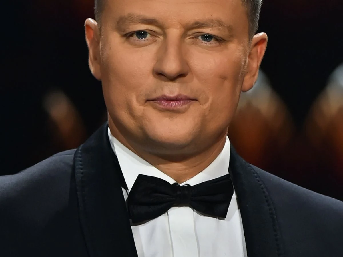 Rafał Brzozowski Mężczyzna w smokingu i muszce, stojący na tle o ciemnym odcieniu.