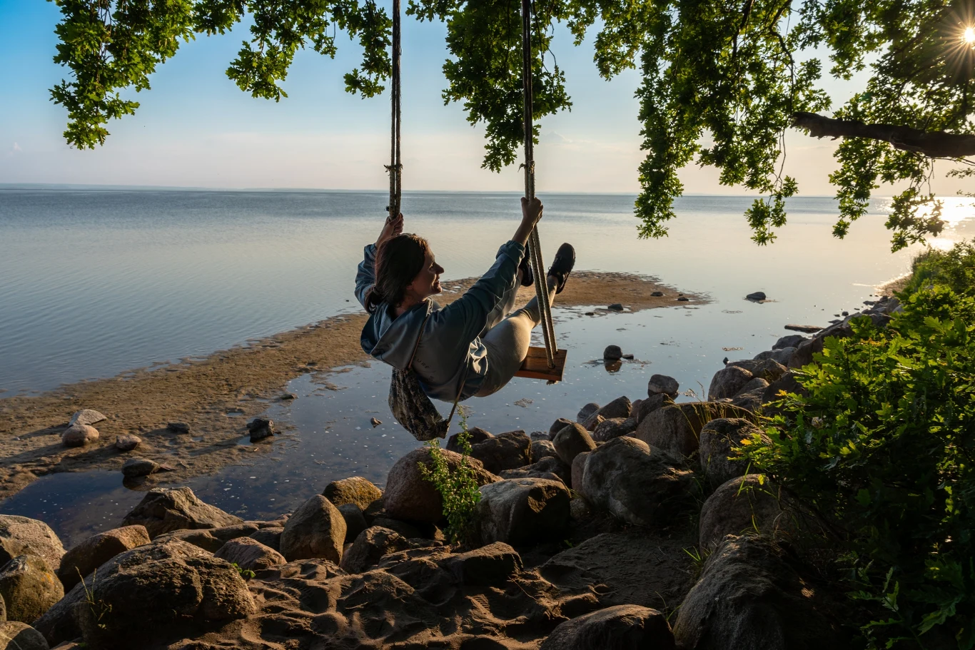 Osoba siedząca na huśtawce zawieszonej na drzewie nad brzegiem jeziora, otoczona dużymi kamieniami i zielenią, z widokiem na spokojną wodę i nisko świecące słońce.