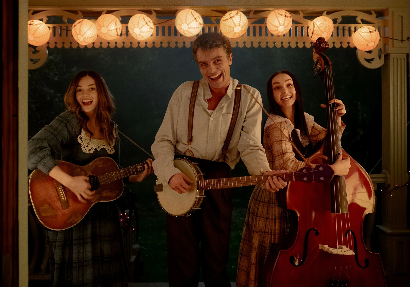 Trójka młodych muzyków w strojach retro grających na gitarze, banjo i kontrabasie, uśmiechniętych i stojących pod jasnymi lampionami na werandzie.