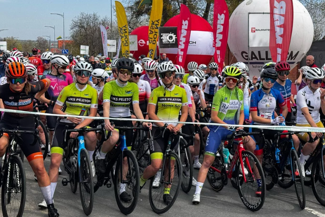 Grupa zawodniczek na rowerach szosowych w kolorowych strojach kolarskich, ustawionych na linii startu przed wyścigiem, w otoczeniu licznych flag sponsorów i dużego balonu reklamowego.