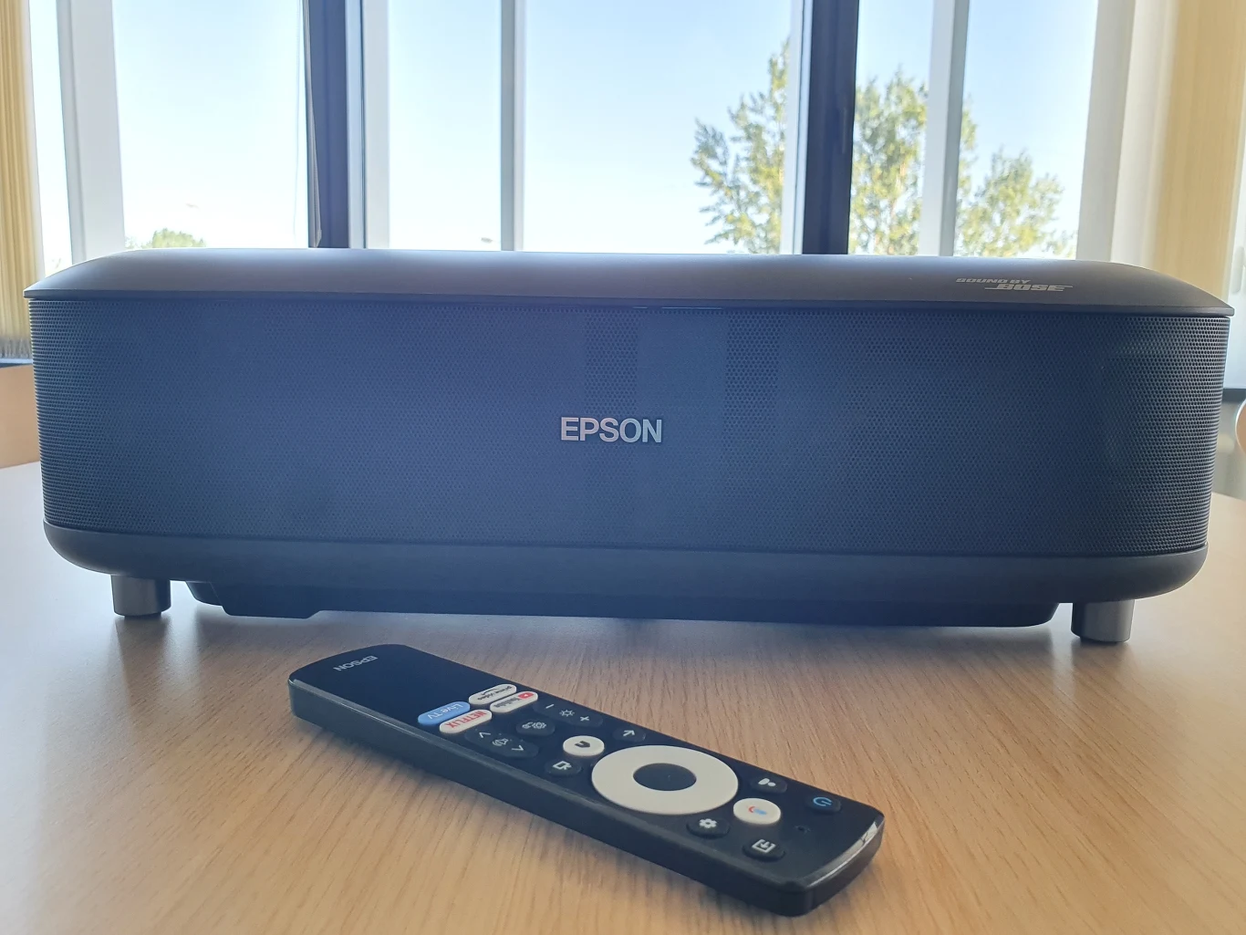 Lifestudio Grand EH-LS670 Czarny projektor Epson ustawiony na drewnianym stole, przed nim leży pilot z wyraźnymi przyciskami funkcyjnymi.