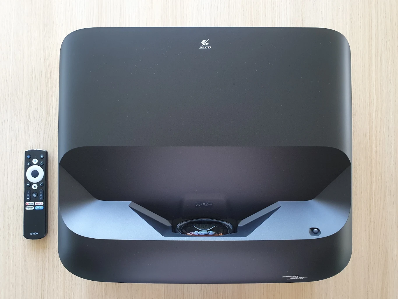 Lifestudio Grand EH-LS670 Nowoczesny projektor o czarnej, prostokątnej obudowie umieszczony na jasnym blacie, obok którego leży czarny pilot z białymi przyciskami.