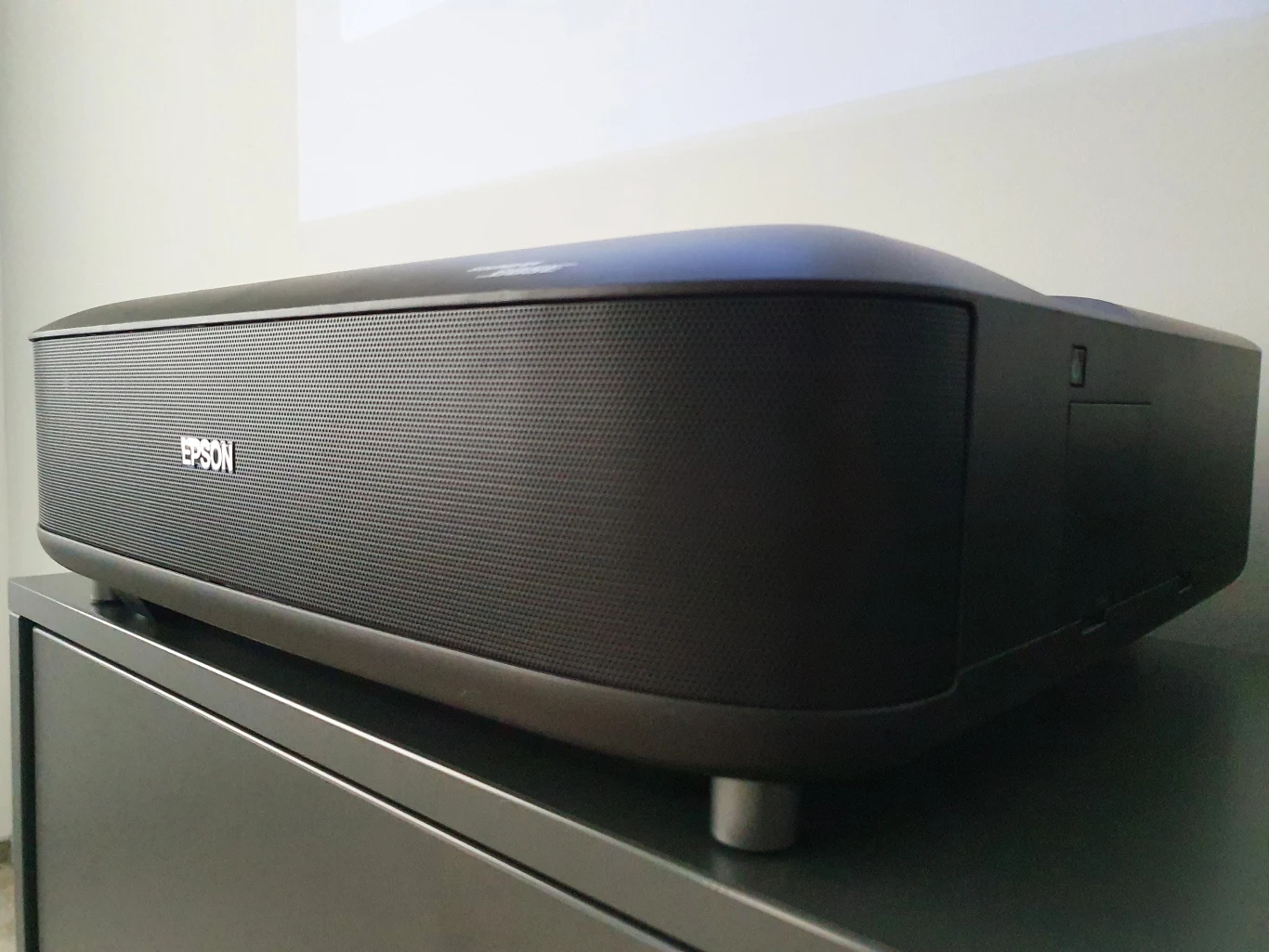 Lifestudio Grand EH-LS670 Czarny projektor marki Epson ustawiony na metalowej szafce, skierowany w stronę białej ściany.