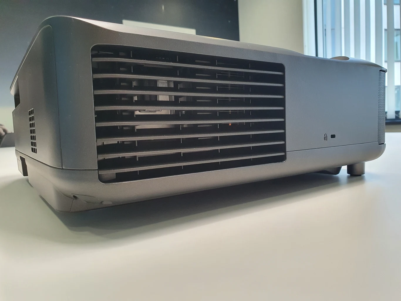 Lifestudio Grand EH-LS670 Szary projektor multimedialny ustawiony na białym blacie, z widoczną kratką wentylacyjną na boku oraz dużym oknem w tle po prawej stronie.