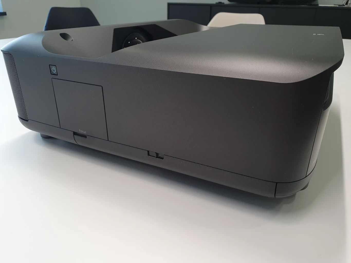 Lifestudio Grand EH-LS670 Czarny projektor ustawiony na jasnym stole w nowoczesnym biurze, w tle krzesła i szafka.