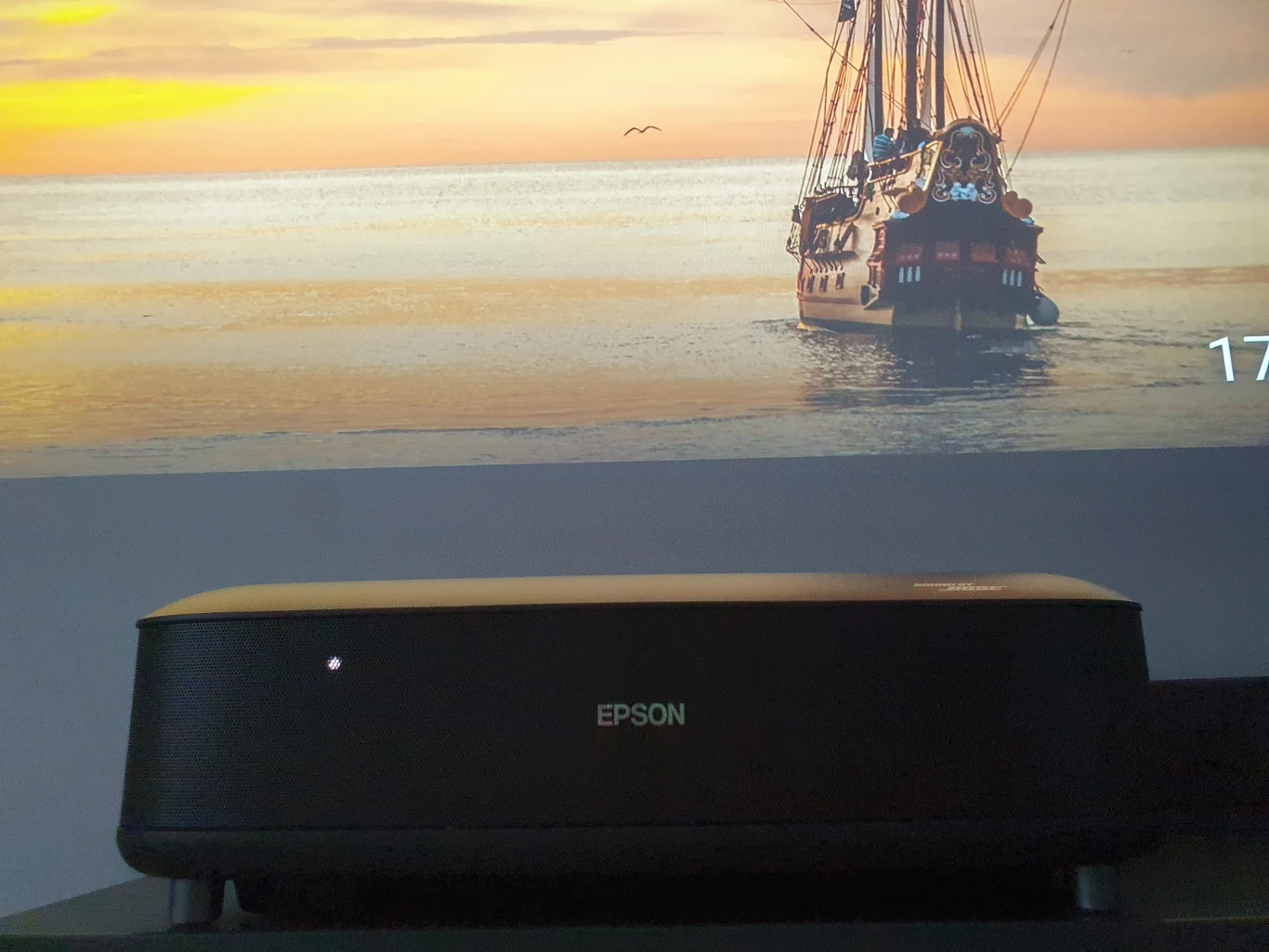 Lifestudio Grand EH-LS670 Projektor Epson ustawiony na ciemnej powierzchni, wyświetlający na ścianie obraz żaglowca płynącego po spokojnym morzu o zachodzie słońca.
