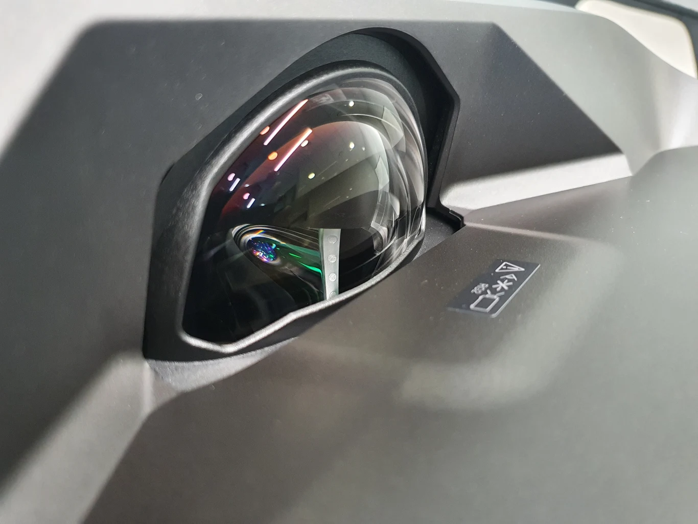 Lifestudio Grand EH-LS670 Duża soczewka projektora Head-Up Display wbudowana w deskę rozdzielczą samochodu, z wyraźnymi odbiciami światła na szklanej powierzchni oraz widoczną ikoną oznaczenia na czarnym panelu obok.