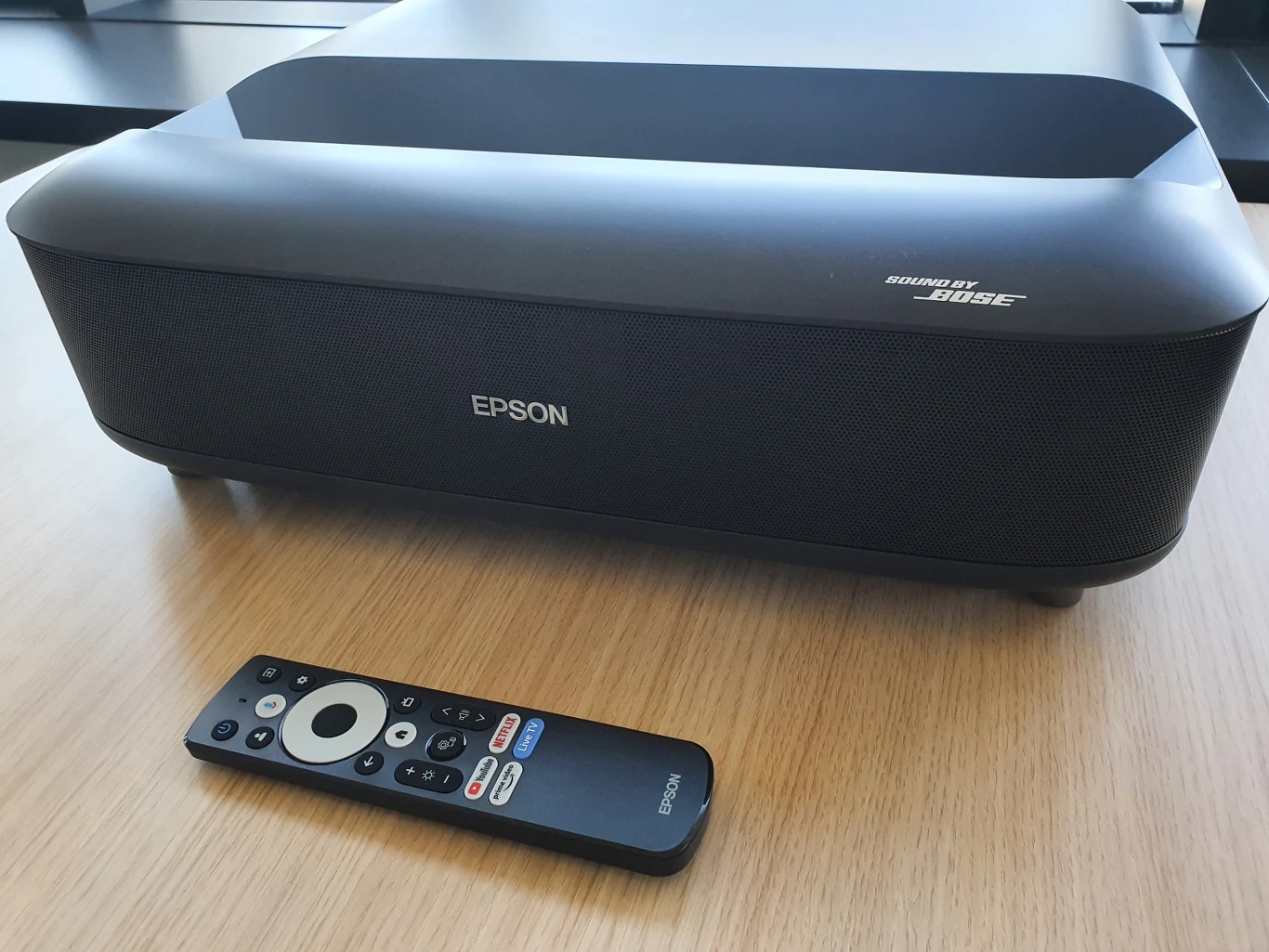 Lifestudio Grand EH-LS670 Projektor Epson z nagłośnieniem sygnowanym przez Bose oraz dedykowany pilot leżący na jasnym drewnianym blacie.