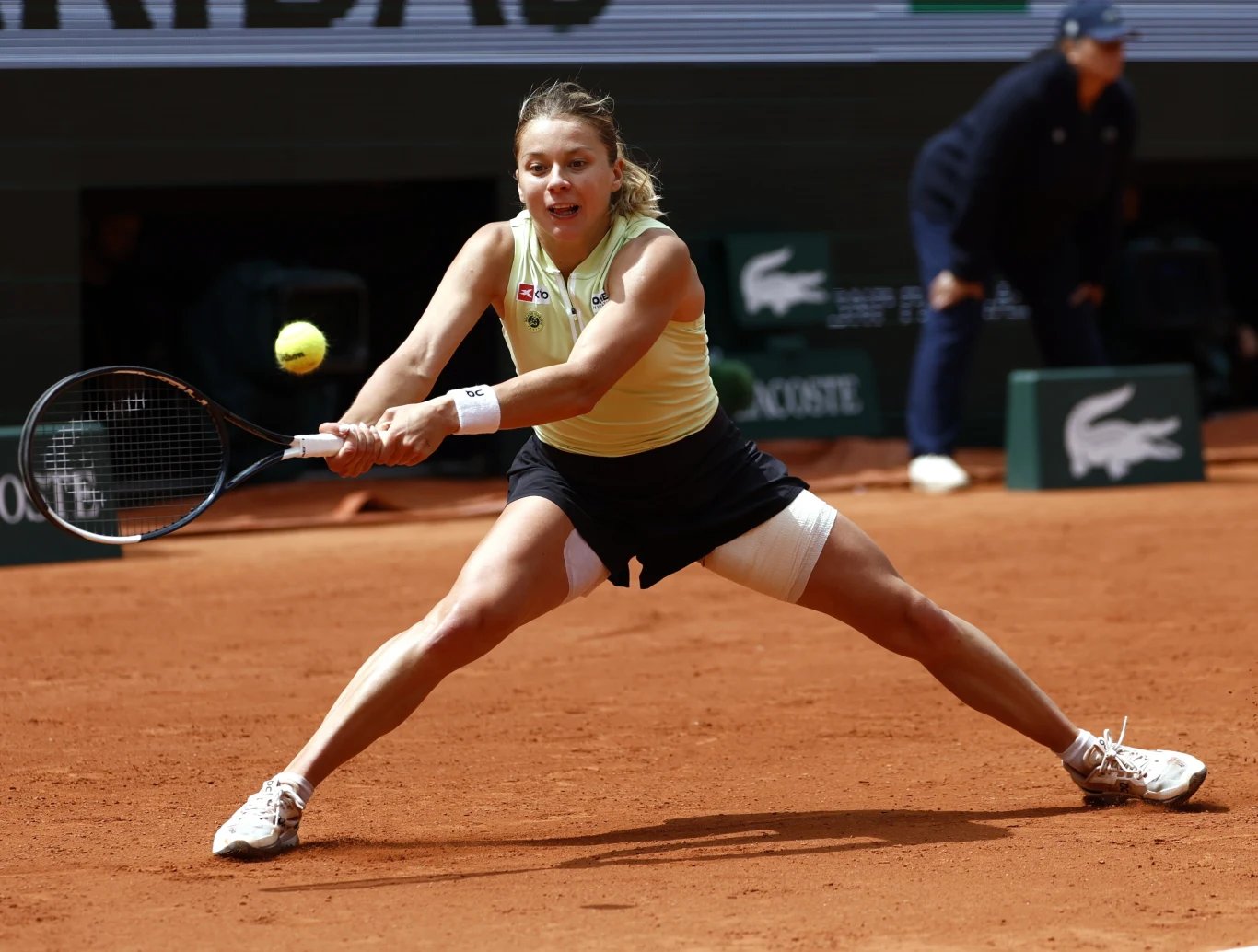Tenisistka w sportowym stroju wykonuje szeroki rozkrok, uderzając rakietą piłkę na kortcie ziemnym podczas meczu.