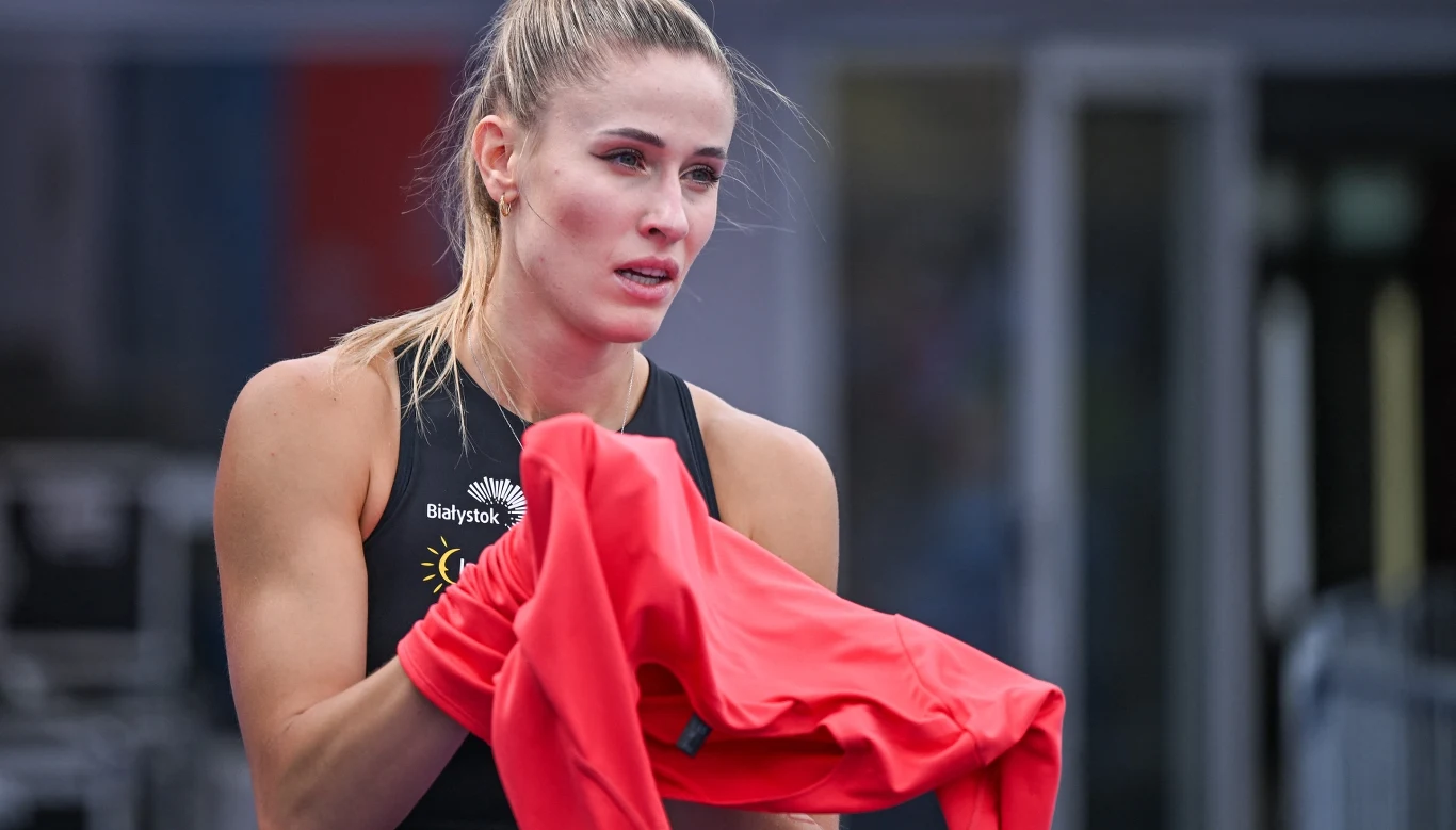 Kobieta w sportowym stroju trzyma w rękach czerwoną kurtkę, ma spięte włosy i skupiony wyraz twarzy.