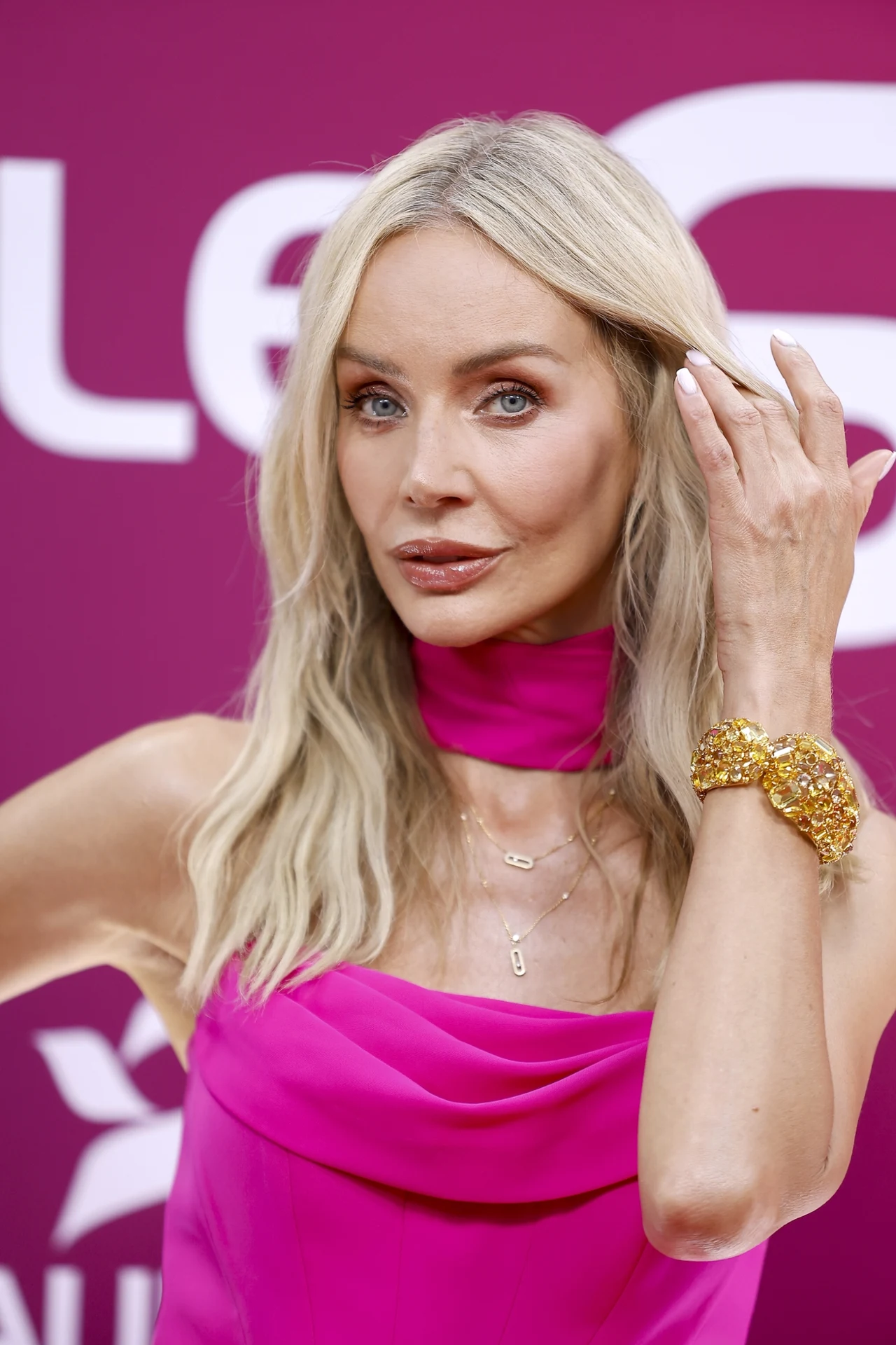 Kobieta o długich blond włosach w różowej sukni z dopasowaną apaszką, ozdobna złota bransoletka na nadgarstku, neutralny makijaż i spokojny wyraz twarzy na tle w odcieniach fioletu.