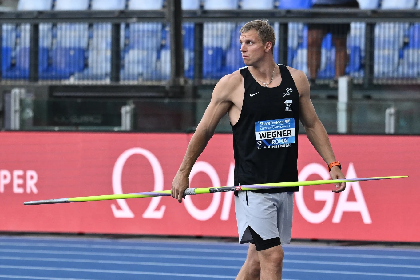 Lekkoatleta w sportowym stroju trzymający oszczep na tle bieżni stadionu lekkoatletycznego.