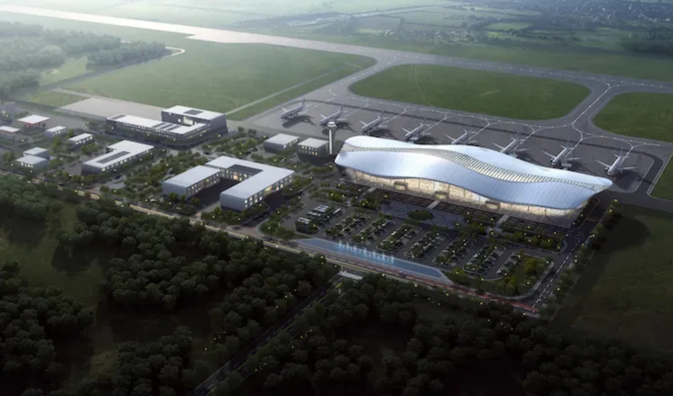 Nowoczesny terminal lotniczy z falistym dachem, kilka zaparkowanych samolotów przy rękawach, kompleks budynków obsługujących lotnisko oraz parking otoczony zielenią.