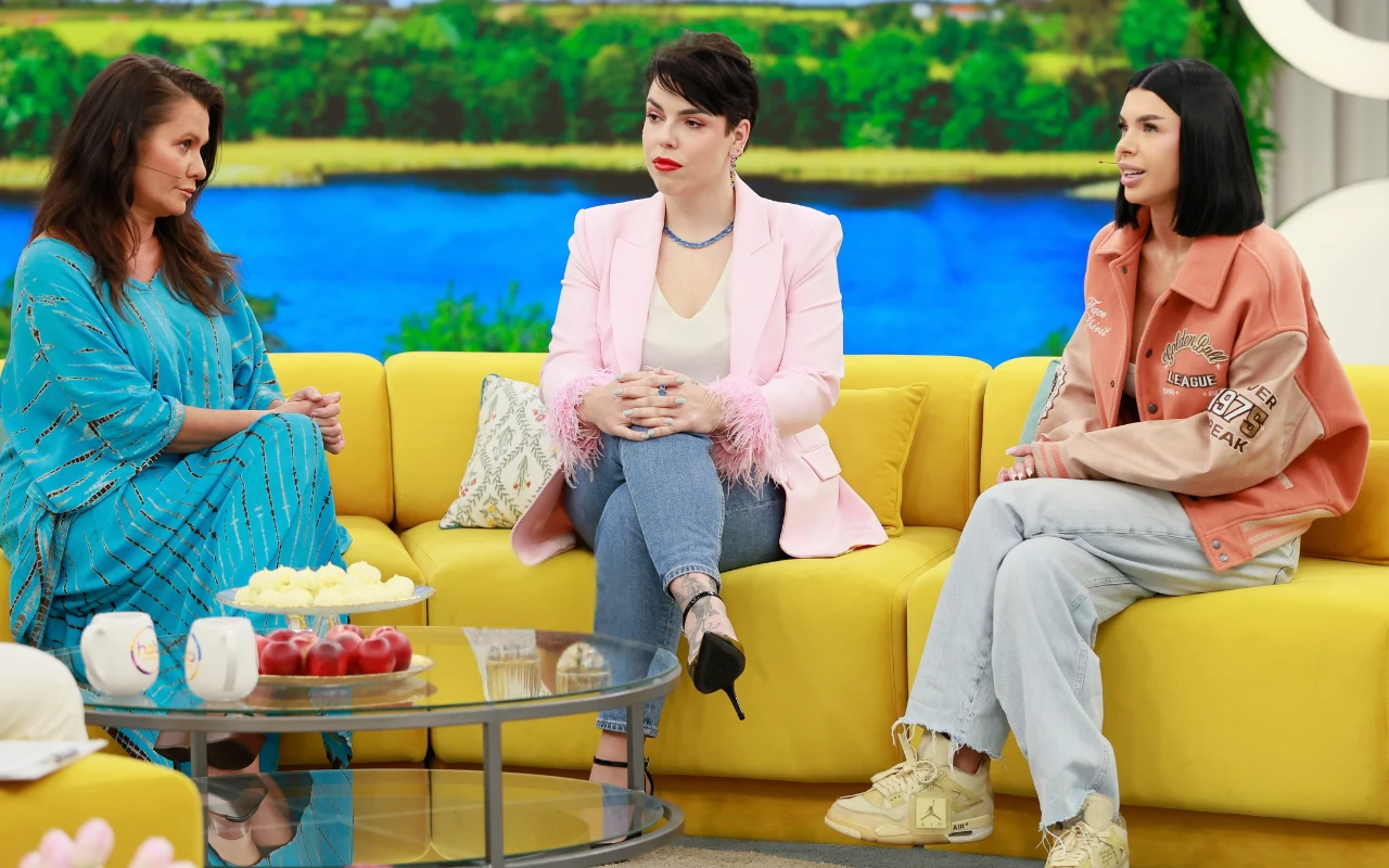 Trzy kobiety siedzące na żółtej kanapie w studiu telewizyjnym, za nimi panoramiczne okno z widokiem na zieleń i wodę, przed nimi przezroczysty stolik z owocami i filiżankami.