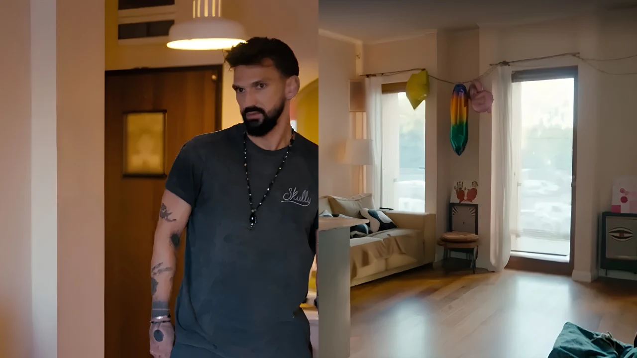 Tak mieszka Jacek z "Love is Blind: Polska" Mężczyzna z brodą i tatuażami stoi w jasnym mieszkaniu z sofą, oknami balkonowymi i kolorowymi dekoracjami zawieszonymi na ścianie.