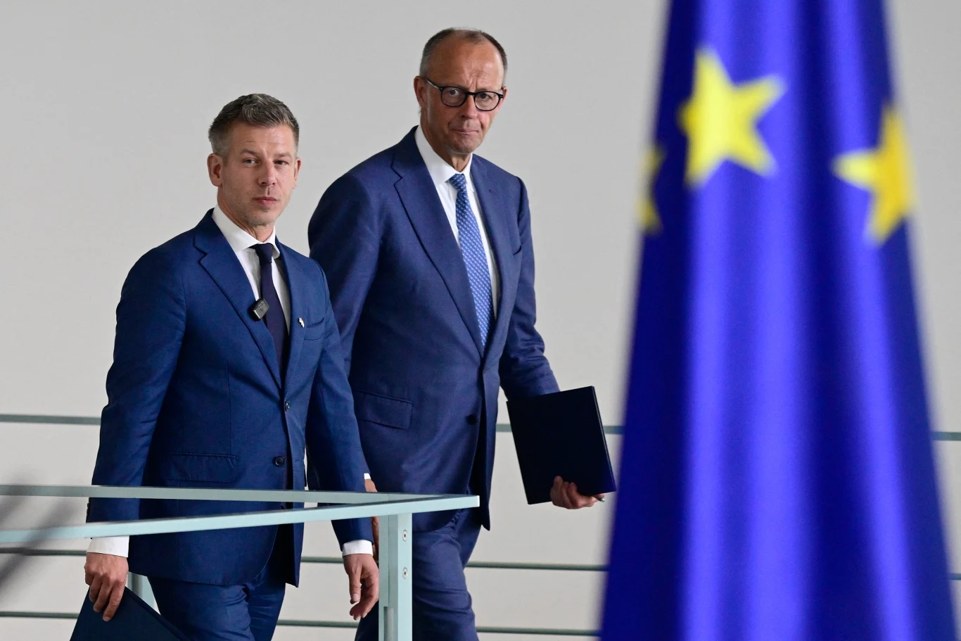 Dwóch elegancko ubranych mężczyzn w garniturach trzymających dokumenty, idących w stronę widocznej z przodu flagi Unii Europejskiej.
