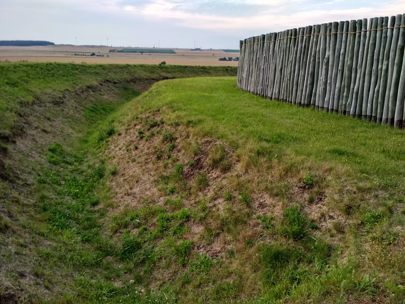 Rekonstrukcja rondela w niemieckim Goseck. Te w Polsce wyglądały bardzo podobnie, fot. S. Zdziebłowski drewniana palisada osadzona na wzniesieniu z szerokim wałem ziemnym, otoczona zieloną trawą, w tle rozległe pola uprawne