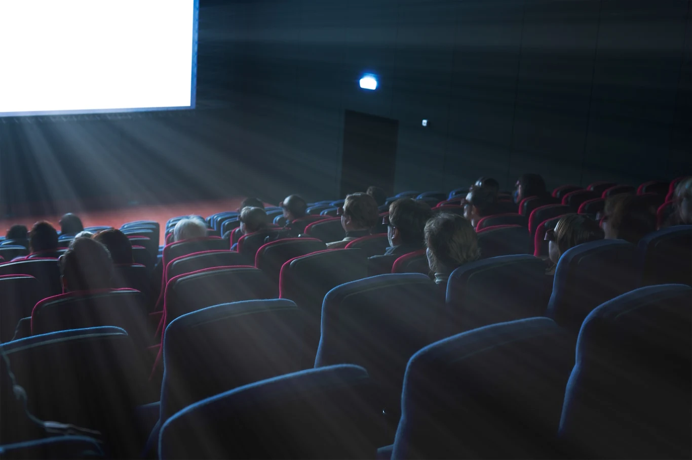 Widownia w kinie skupiona na oglądaniu filmu, w promieniach światła wydobywających się z projektora na dużym ekranie.