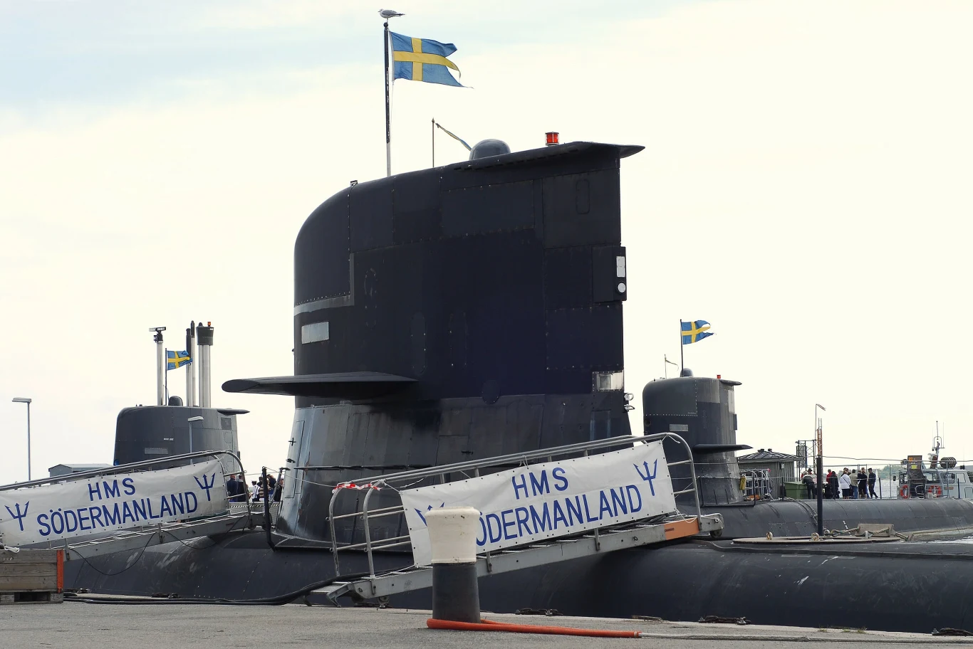 Szwedzki okręt podwodny HMS Södermanland z wyraźnie widocznymi kioskiem i banderą Szwecji, zacumowany przy nabrzeżu portowym.